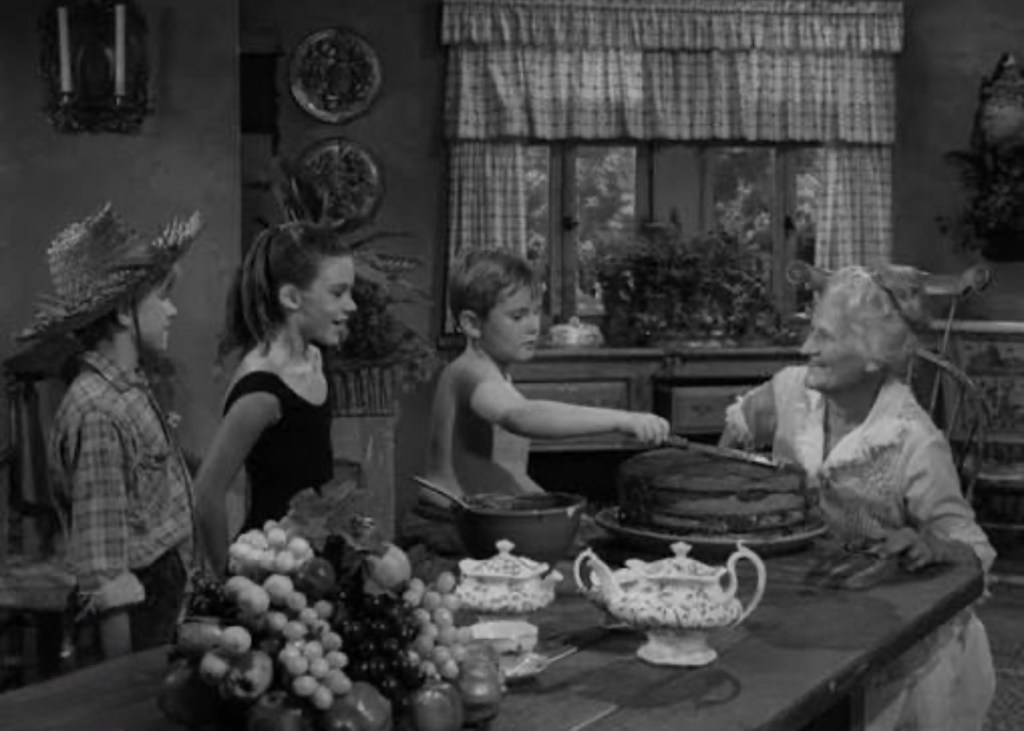 Children interacting with an elderly woman in a kitchen, with a large cake on the table and a variety of fruits in the foreground.