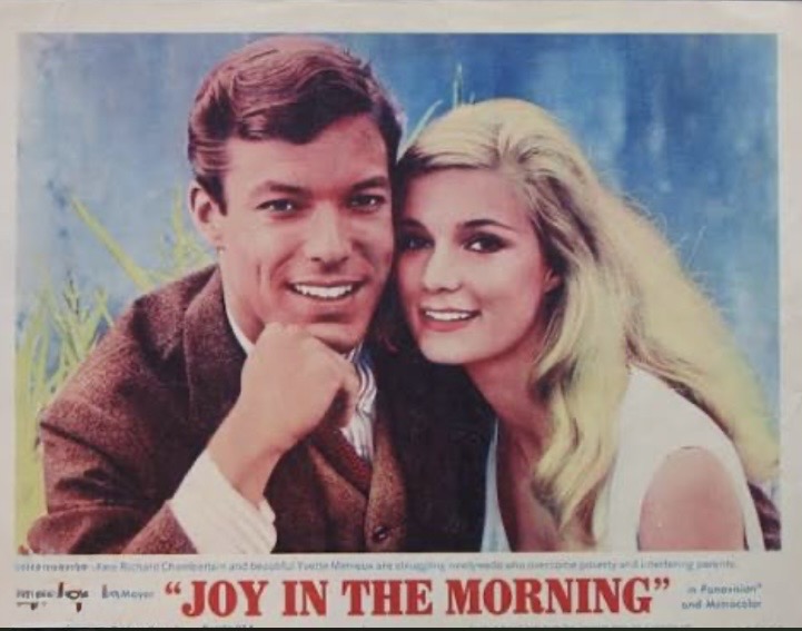 A smiling couple posing together against a light blue background, featuring a man in a brown jacket and a woman with long blonde hair in a white dress. The image is promotional for the film 'Joy in the Morning.'