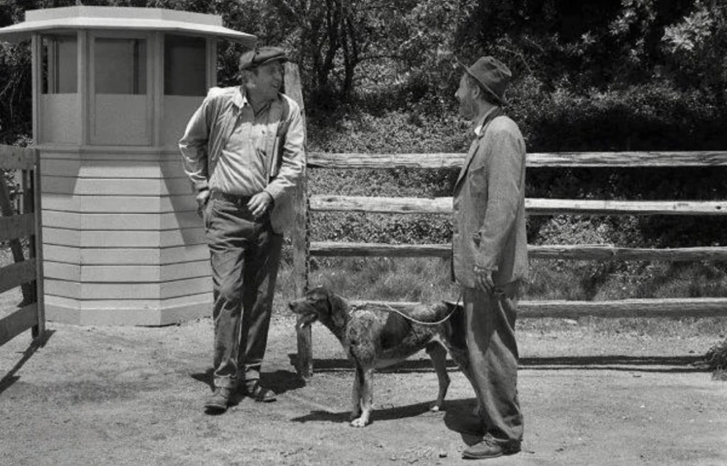 Two men standing beside a fenced area, one holding a leash attached to a dog. In the background, there is a small watchtower and trees.