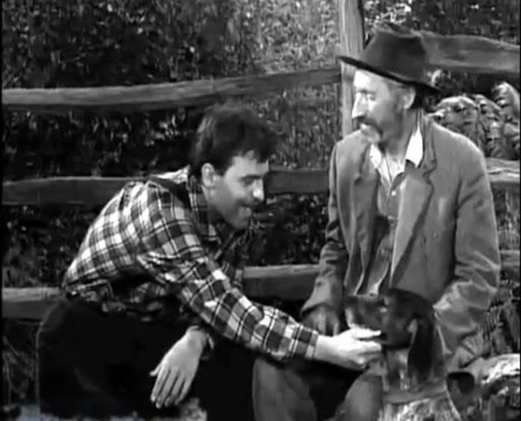 A man in a plaid shirt interacts with a dog while sitting next to another man in rustic attire, all set against a wooden fence.
