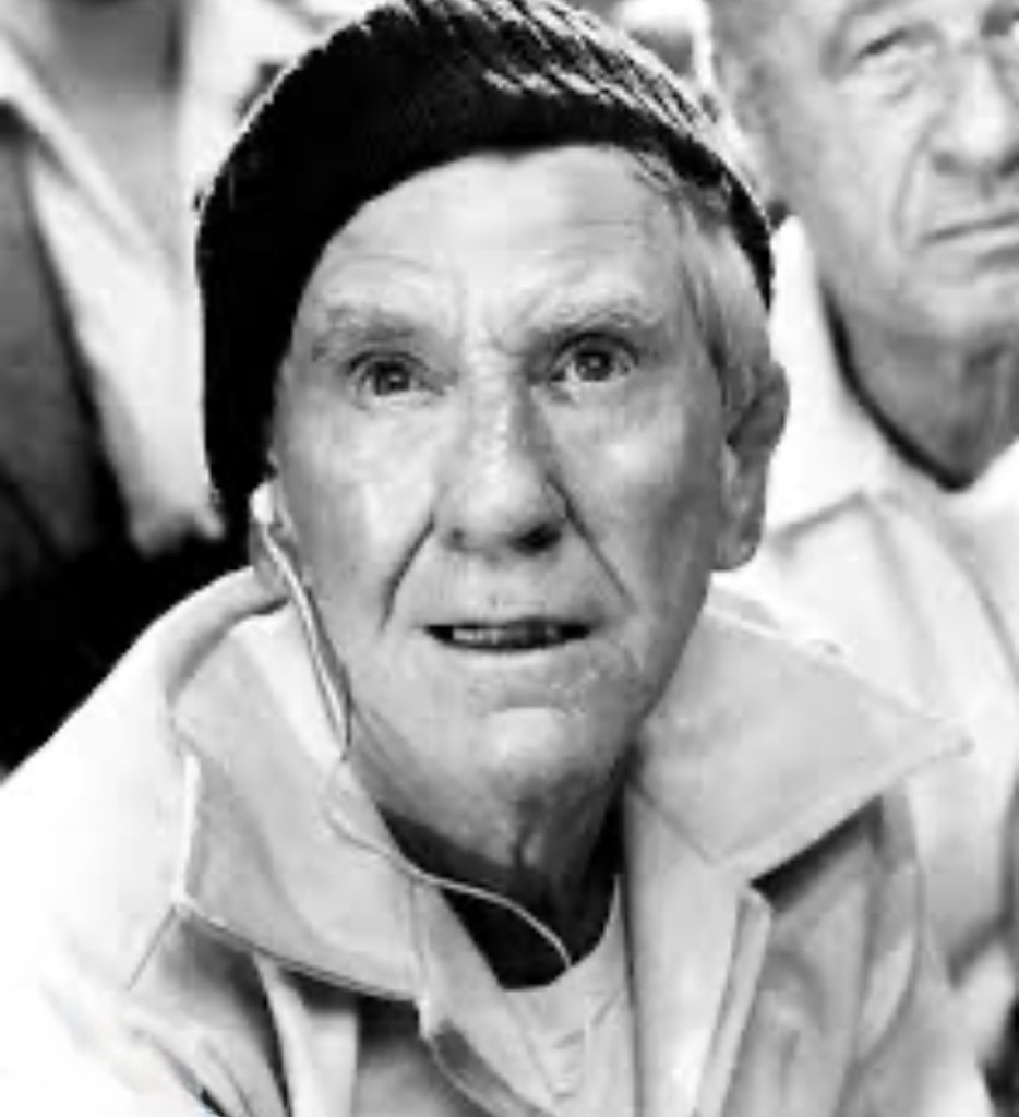 Close-up portrait of an elderly man wearing a black beret and a light-colored jacket, looking directly at the camera with a serious expression.