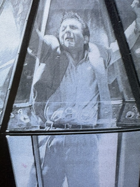 A man inside a glass enclosure, exhibiting a distressed expression while struggling to break free from the restraints.