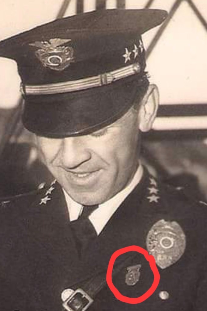 A historic black and white photo of a smiling police officer wearing a cap with insignia and a uniform adorned with badges, focusing on a specific badge highlighted in a red circle.
