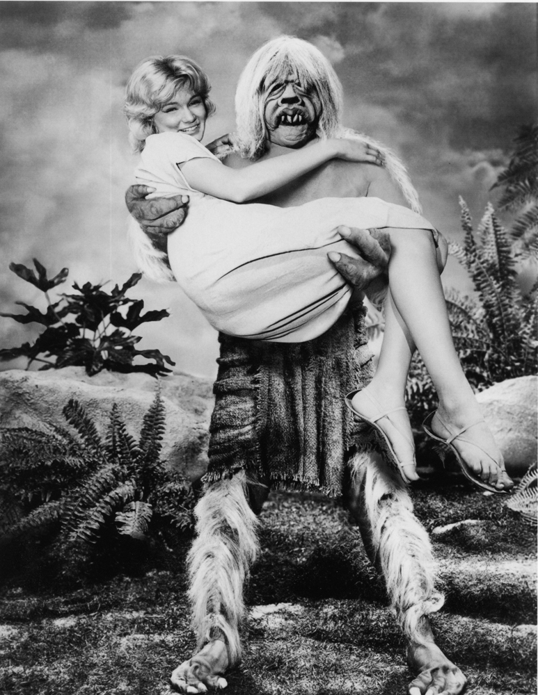 A mythical creature with long hair and animal-like features is holding a woman in a white dress, smiling, in a lush, prehistoric setting.