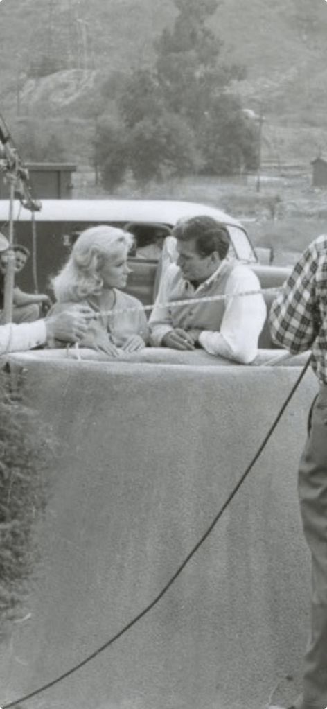 Black and white image of a film set with a woman and man sitting on a ledge engaged in conversation, while crew members are visible in the background adjusting equipment.