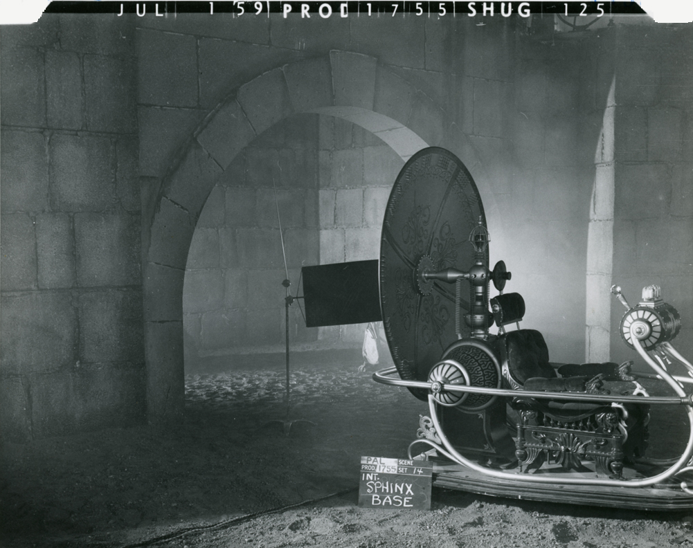 A vintage film set featuring an elaborate, futuristic vehicle against a backdrop of stone walls. The scene is dimly lit with a misty atmosphere, and there is a sign labeled 'INT. SPHINX BASE' positioned near the vehicle.