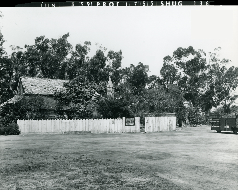 Black and white photograph of a rustic house surrounded by trees, featuring a white picket fence and a 'Private Property, Keep Out' sign.