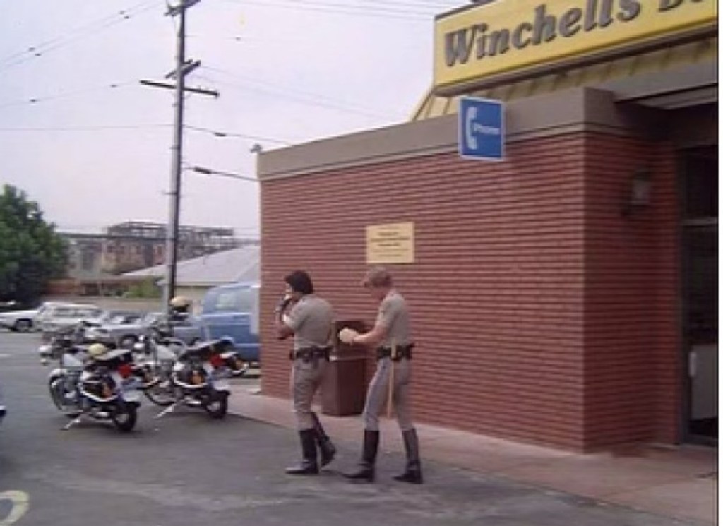 Two police officers walking near a Winchell's Donut shop, with motorcycles parked nearby.