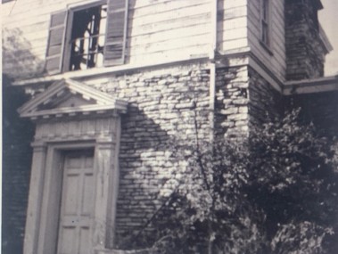 Black and white photograph of an old, two-story building with a stone facade, a wooden door, and partially open shutters on the upper level.