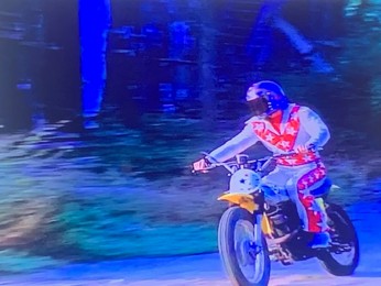 A motocross rider wearing a colorful outfit rides a yellow motorcycle on a dirt path surrounded by trees.