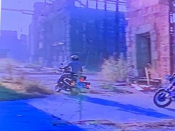 Two motorcyclists riding on a deserted, overgrown street lined with abandoned industrial buildings.