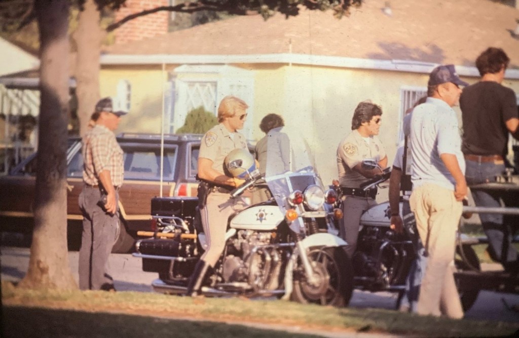 Two motorcycle police officers in uniform standing next to their bikes, with a group of men in casual attire nearby, in front of a house.