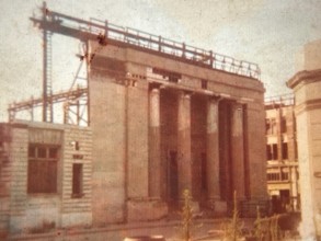 A historical building under construction, featuring prominent columns and scaffolding, with a faded, sepia-toned appearance.