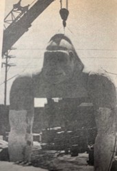 A black and white image of a large gorilla statue in an industrial setting, with a crane in the background and vehicles visible.