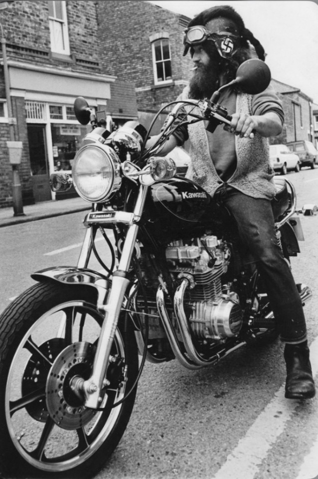 An individual with a beard and goggles, riding a vintage Kawasaki motorcycle on a city street, black and white photography.