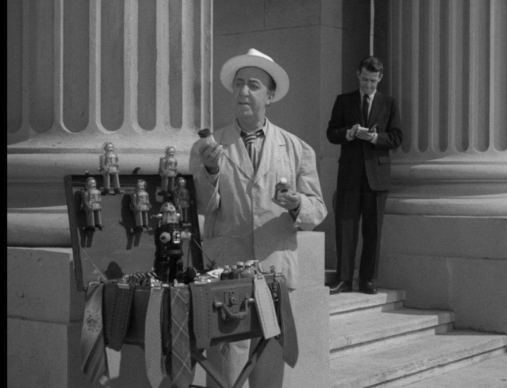 A street vendor in a light-colored coat and white hat displays toys, including robots, from a suitcase in front of a building with columns. A man in a suit stands in the background, taking notes.