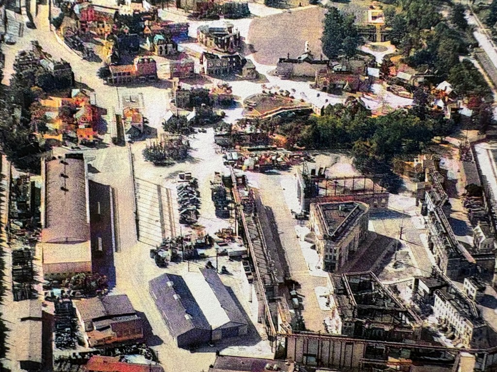 Aerial view of a sprawling construction site with various buildings and structures, including partially completed and demolished areas, surrounded by trees and roads.