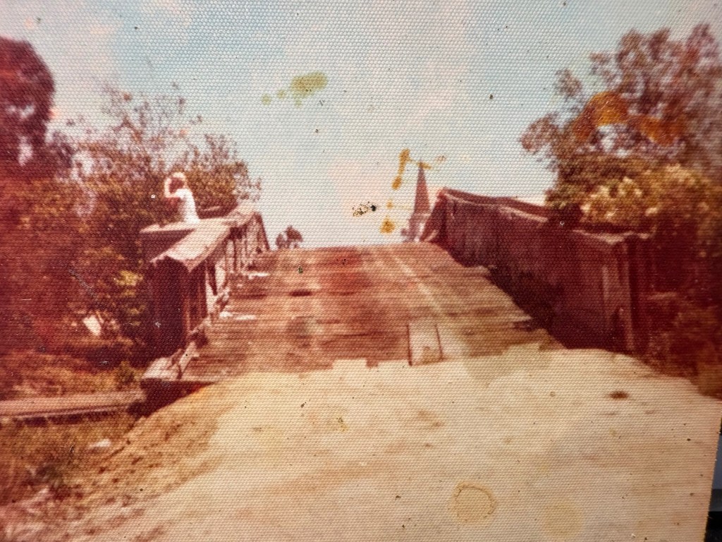 A weathered wooden bridge with visible damage, surrounded by trees and vegetation, under a cloudy sky.