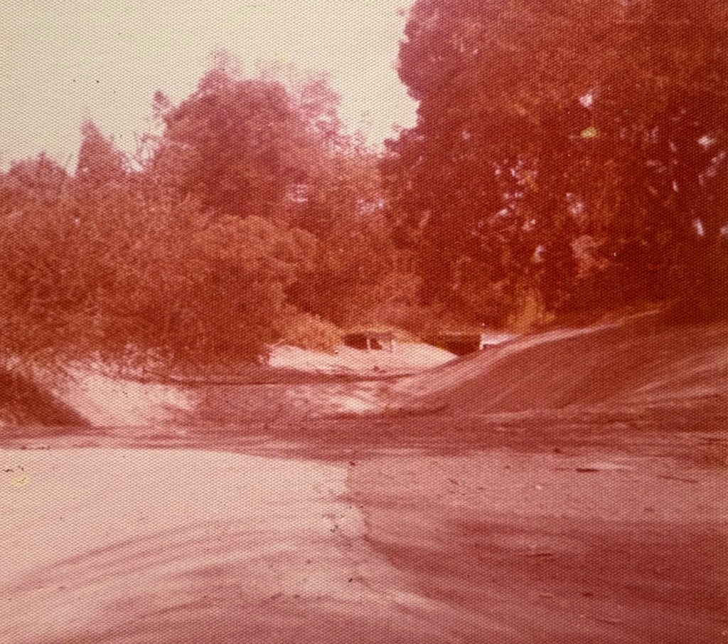 A faded image of a dry riverbed surrounded by trees and bushes, with sandy banks and a clear sky.
