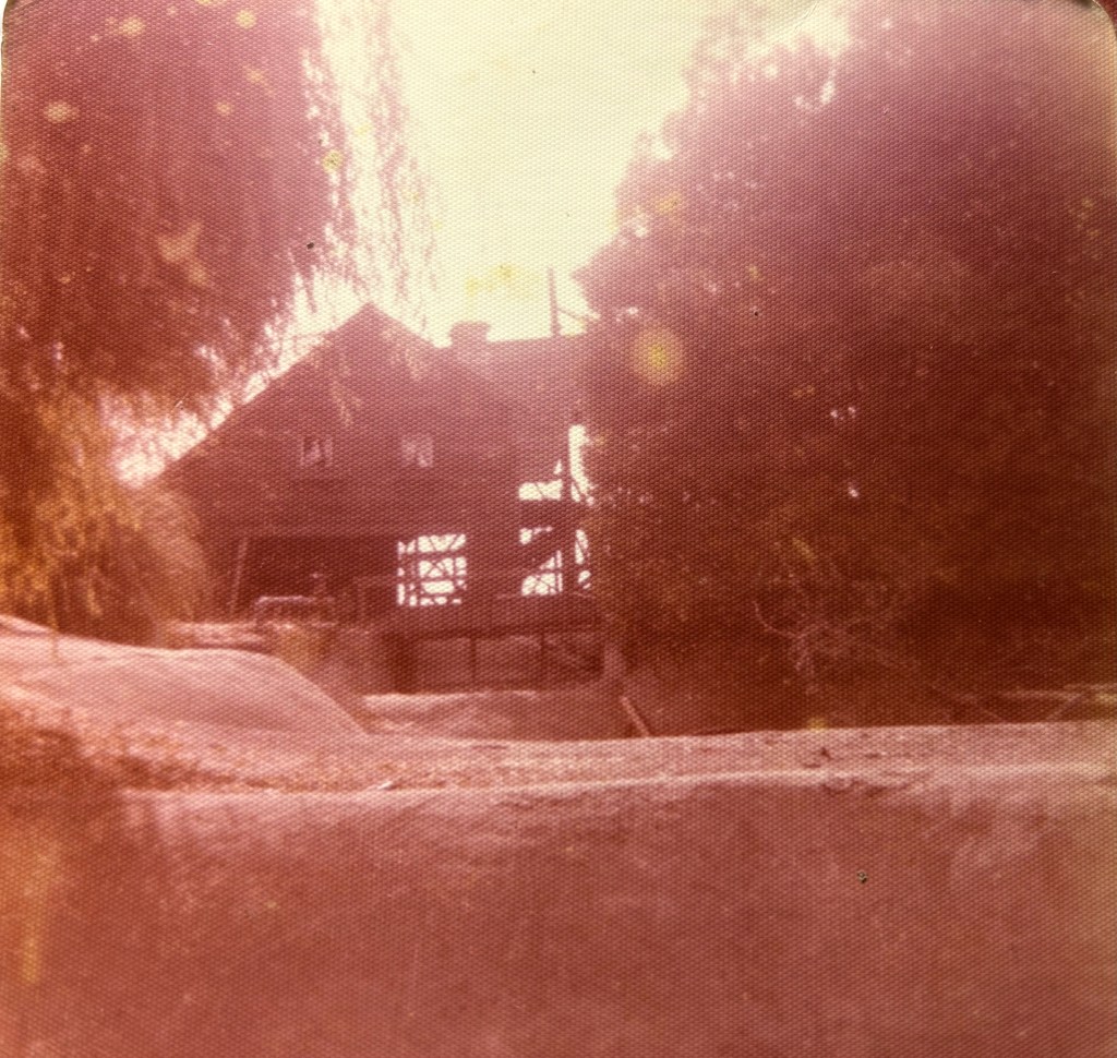 A faded photograph of a rustic house surrounded by trees and natural terrain.