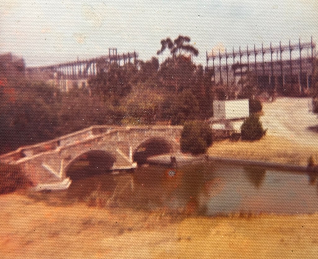 A vintage photograph of a stone bridge arching over a calm pond, surrounded by grassy areas and trees. In the background, a partially constructed building structure is visible.