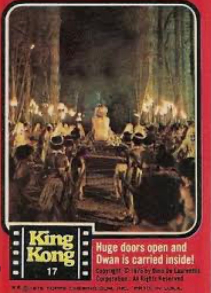 A scene from King Kong featuring a character named Dwan in a dramatic setting with large doors opening, surrounded by onlookers holding torches.