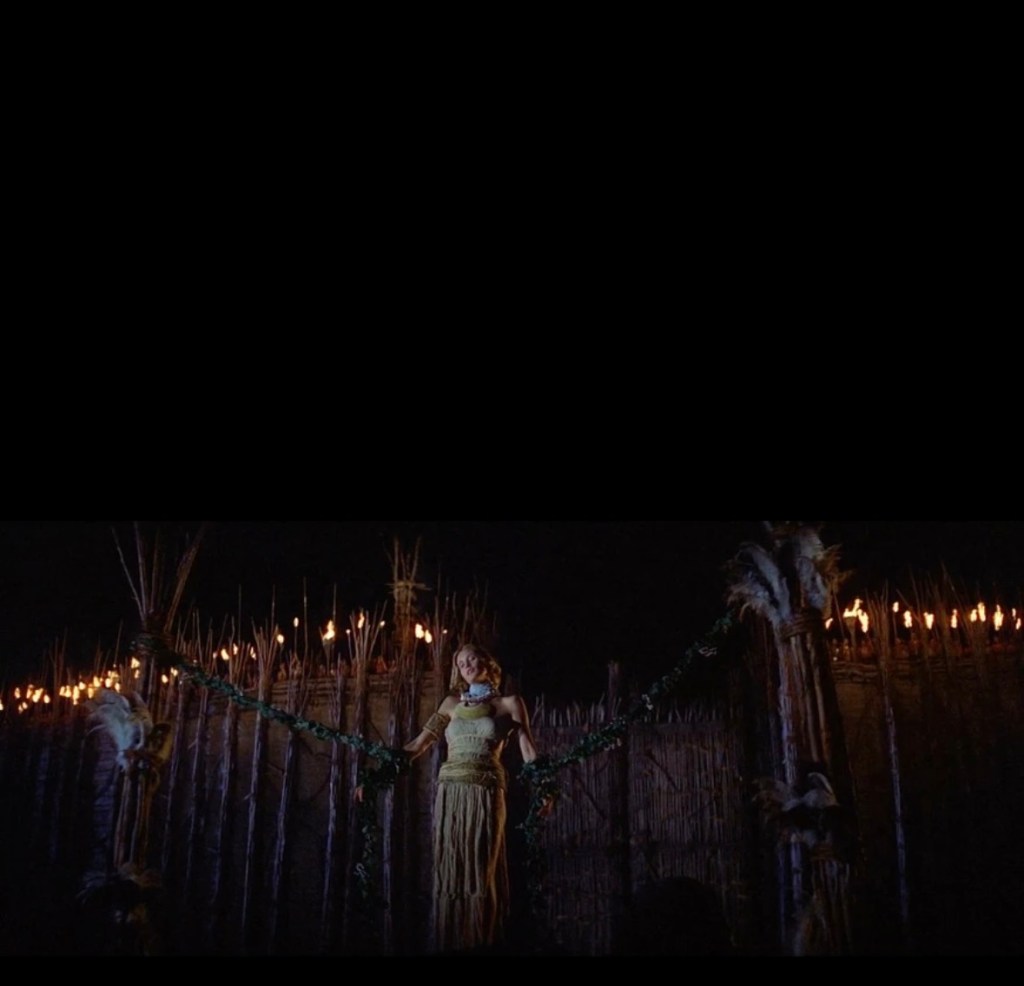A woman dressed in traditional attire stands on a platform adorned with greenery, surrounded by a dark setting illuminated by torches in the background.