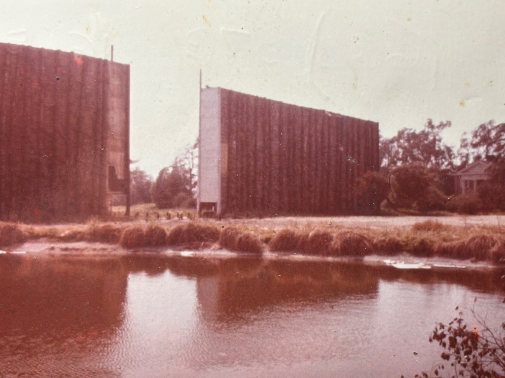 Two large, vertical structures beside a calm water body with grass and trees in the background.
