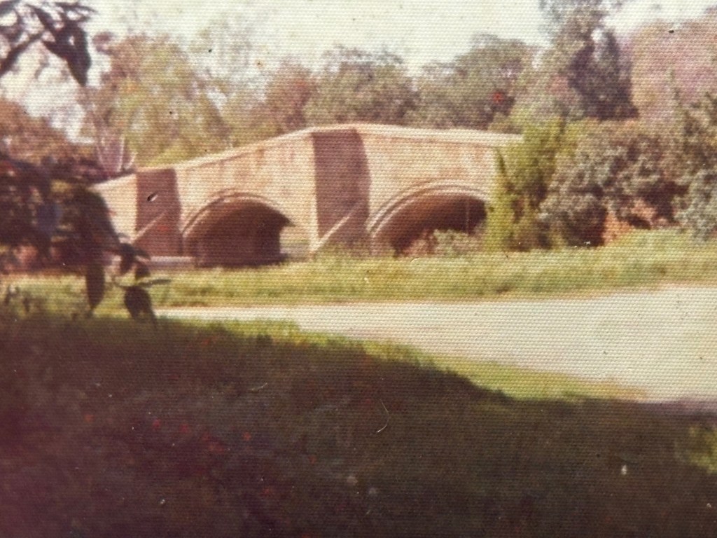 A vintage photograph of a stone bridge with two arches over a calm body of water, surrounded by lush greenery and trees.