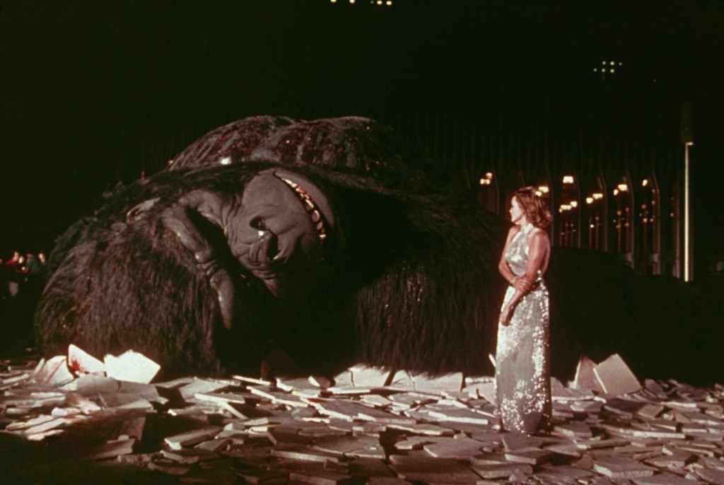 A large gorilla figure lies on the ground with its eyes closed, surrounded by scattered debris, while a woman in a glittering dress stands next to it, looking at the figure.