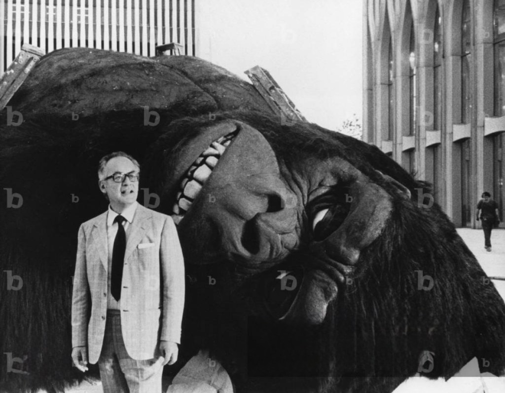 A man in a suit standing next to a large, sculpted gorilla head in an urban setting.