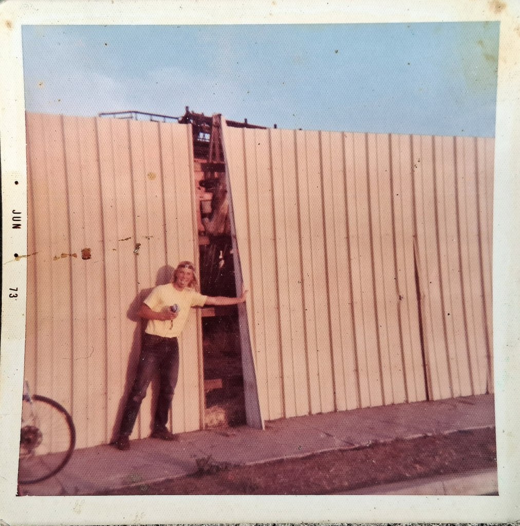 A man in a yellow shirt stands beside a large wooden fence with an opening in it, holding a can. A bicycle is visible in the background.