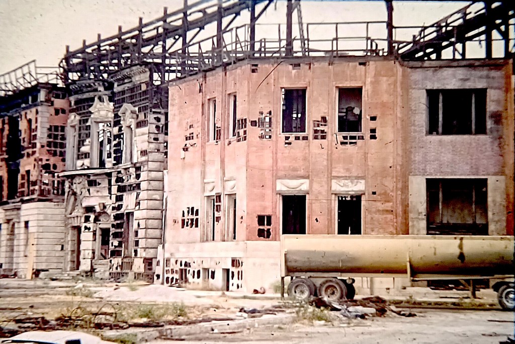 A view of a partially constructed and damaged building with brick and stone facades, showcasing broken windows and exposed structures, alongside a construction vehicle in the foreground.