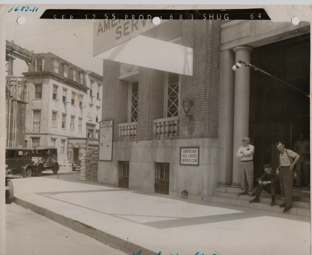 Historical black and white photo of the American Red Cross Service Club, showing people sitting and standing on the steps, with vehicles parked nearby and buildings in the background.