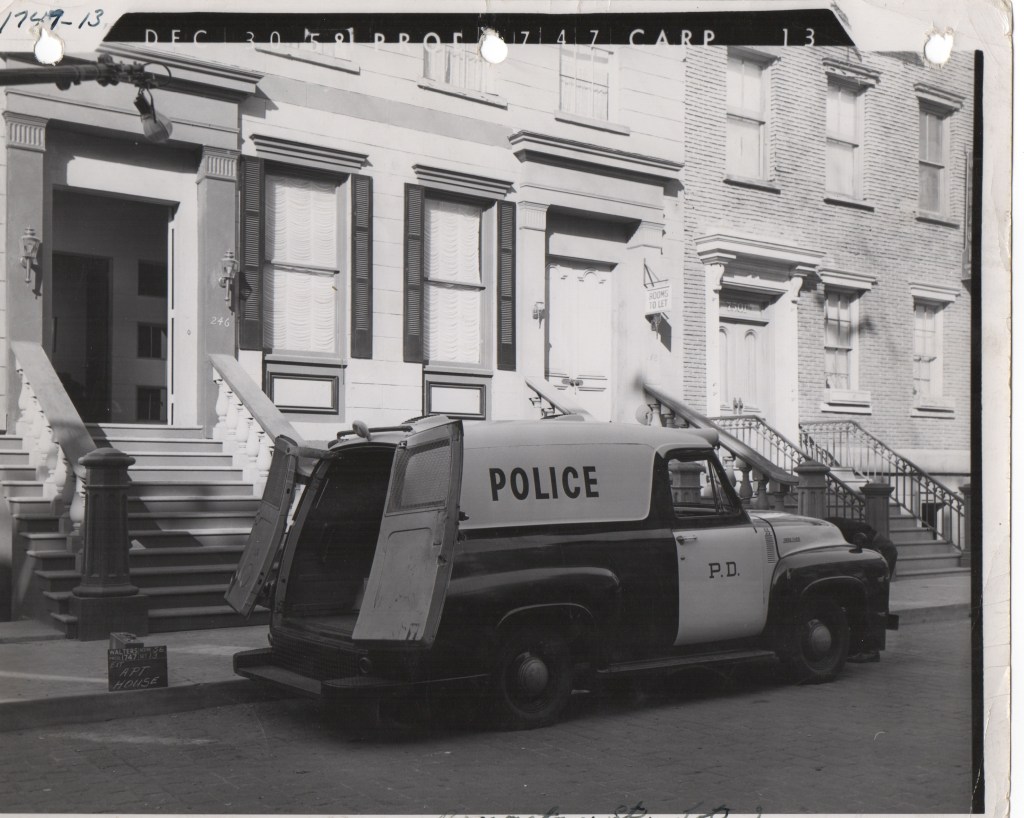 Black and white photo of a vintage police van parked in front of a row of historical buildings with staircases and large windows.