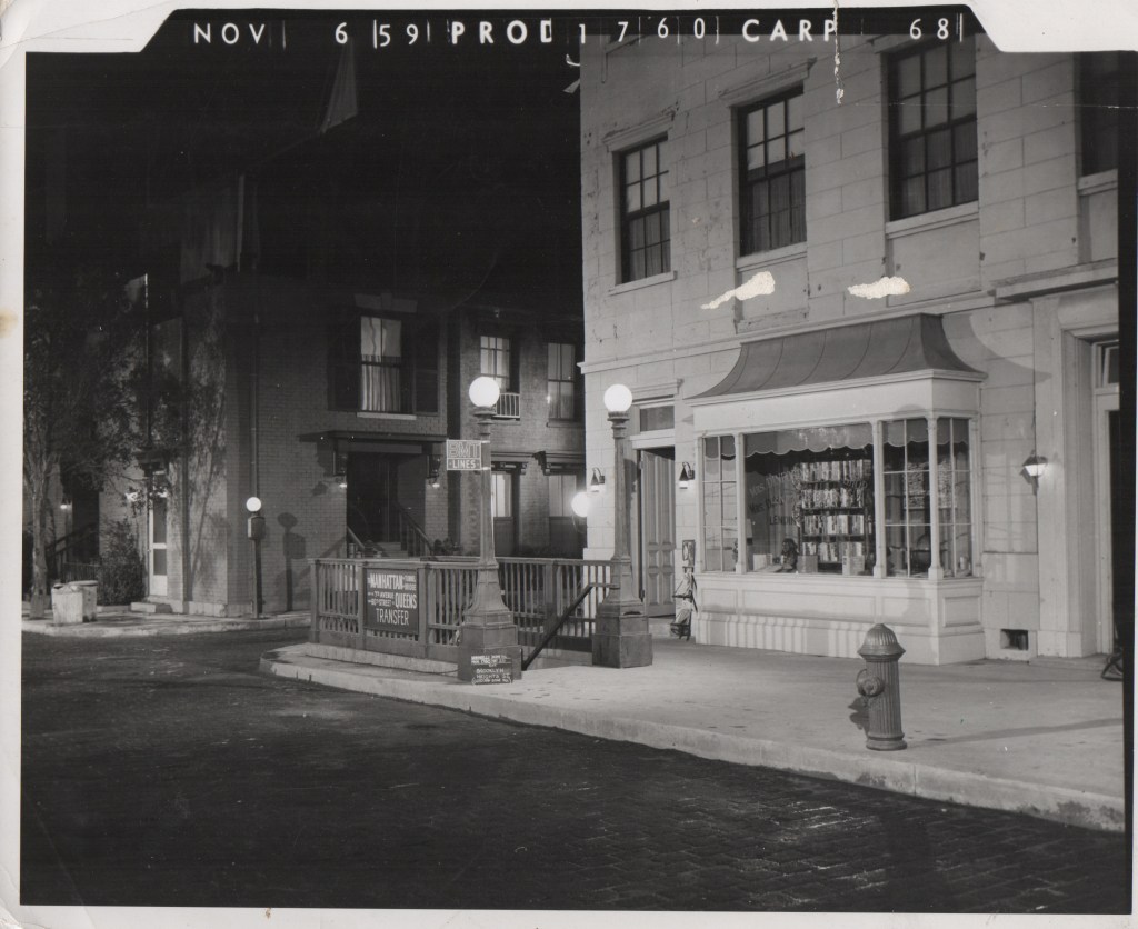 A black and white photograph depicting a street scene at night, featuring a storefront with a display window, street lamps, a fire hydrant, and signs indicating public transport services.