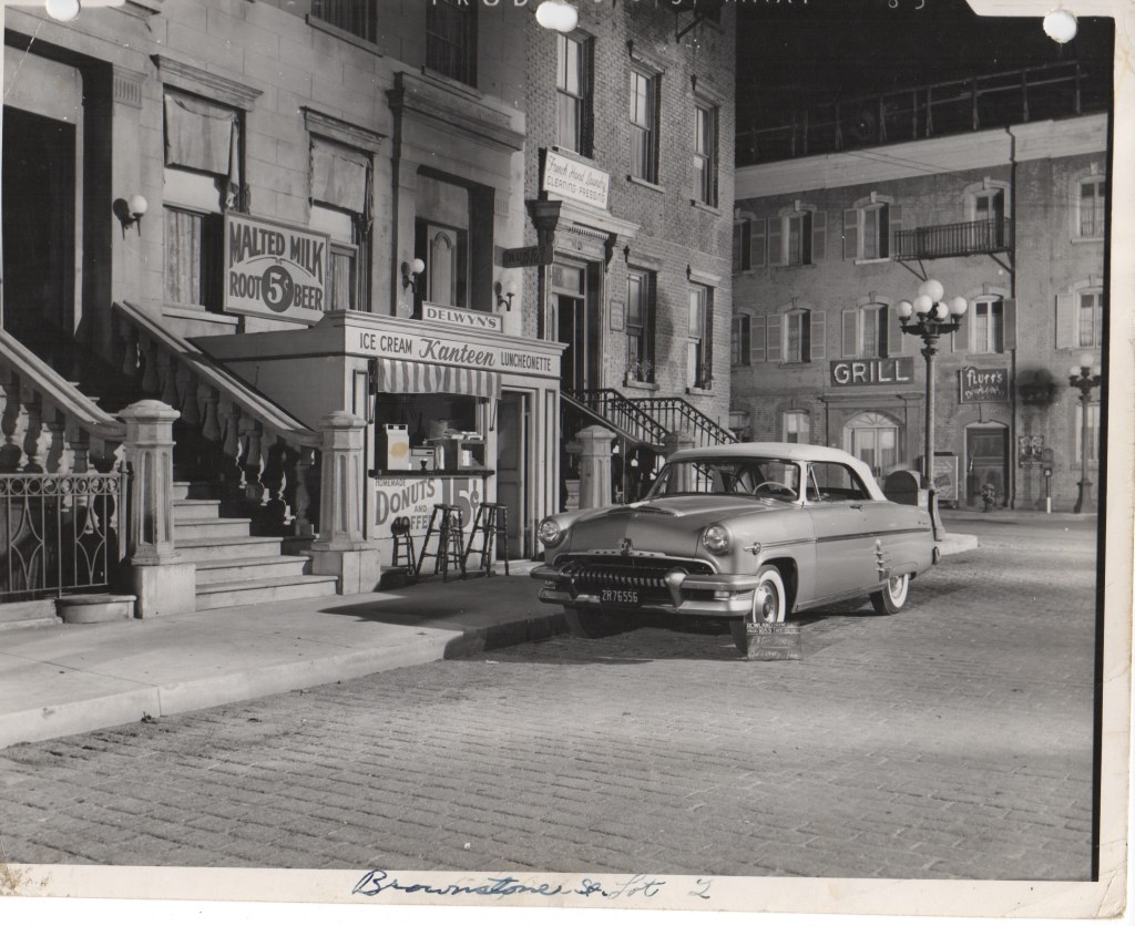 A vintage black and white photograph featuring a classic car parked on a cobblestone street in front of a stylized urban scene, including storefronts for a diner, ice cream parlor, and grill, with ornate street lamps and a historical building backdrop.