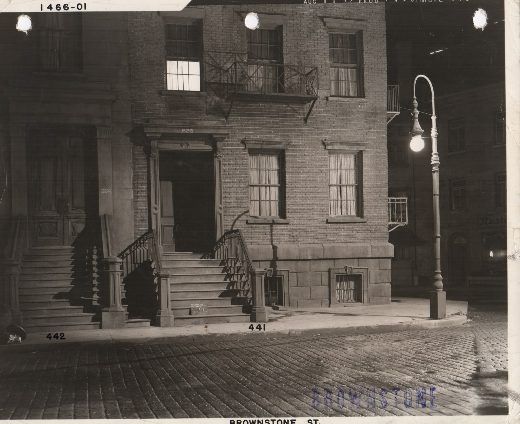 A night view of a brownstone building with stairs leading to the entrance, featuring large windows with curtains. A vintage street lamp is illuminated on the sidewalk, adding a nostalgic atmosphere to the scene.