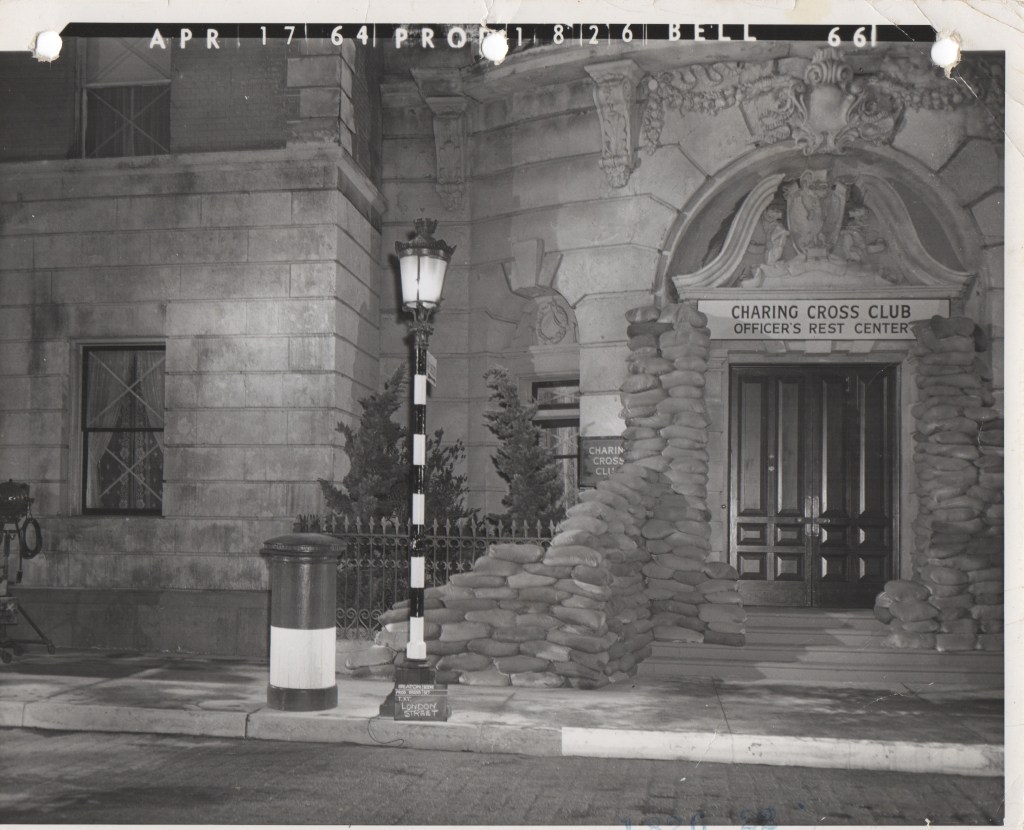 Black and white photograph of the Charing Cross Club entrance, featuring sandbags piled around the doorway, a street lamp, and a post box on the sidewalk, dated April 17, 1964.