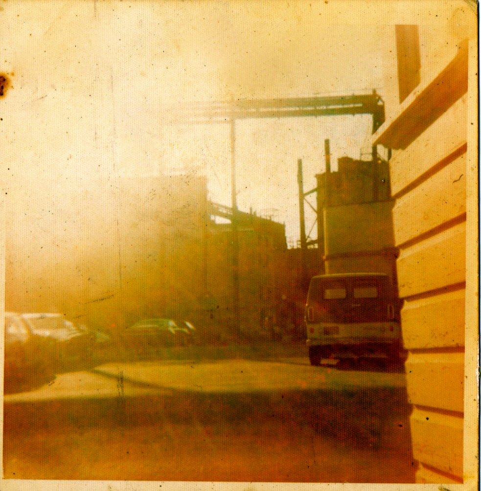 Blurred industrial scene with buildings and parked cars under a hazy sunlight.