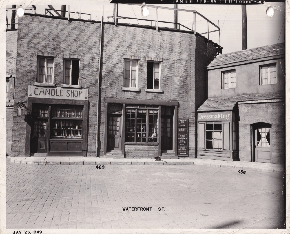 Black and white image of a street scene featuring two buildings: one labeled 'CANDLE SHOP' and the other 'Dressmaking'. The buildings display vintage architecture with detailed brickwork and windows.