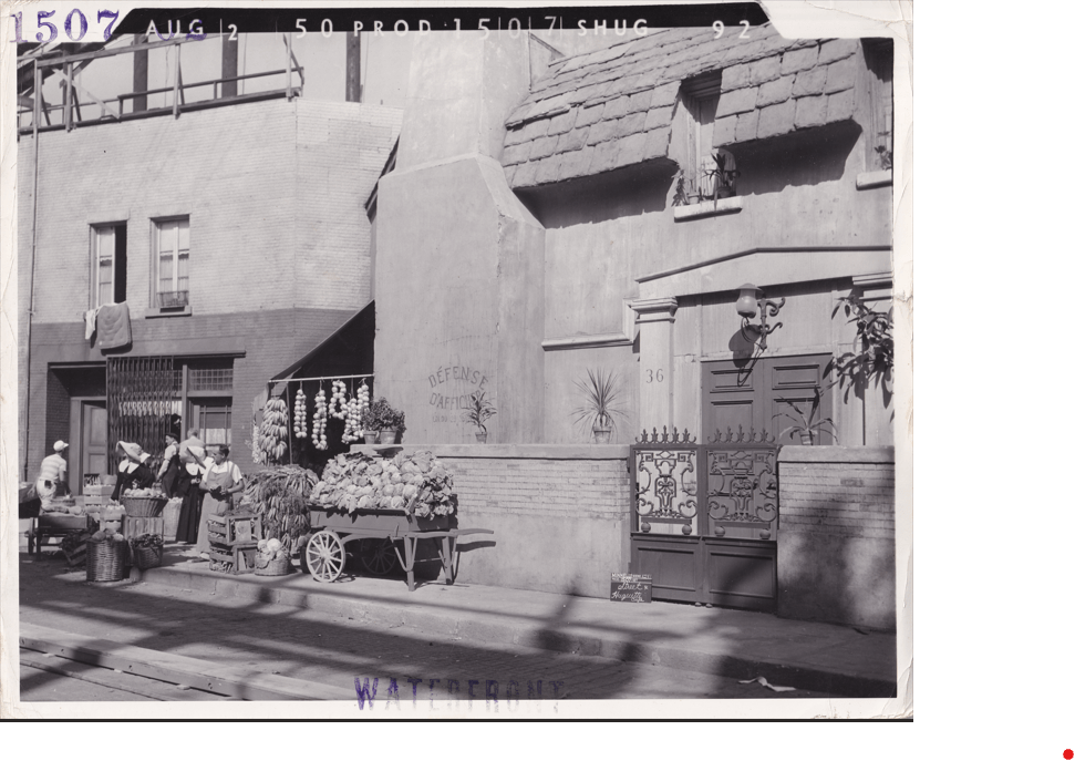 Historical street scene featuring vendors selling fruits and vegetables in baskets, with women dressed in traditional attire. A building with a decorative door is in the background.