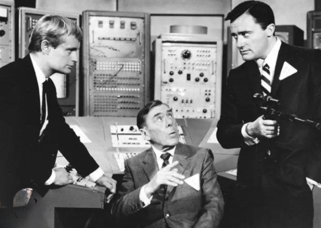 A black and white scene featuring three men in a control room filled with electronic equipment. One man stands to the left wearing a suit and tie, another man is seated in the center looking surprised, and the third man stands to the right holding a gun and wearing a serious expression.
