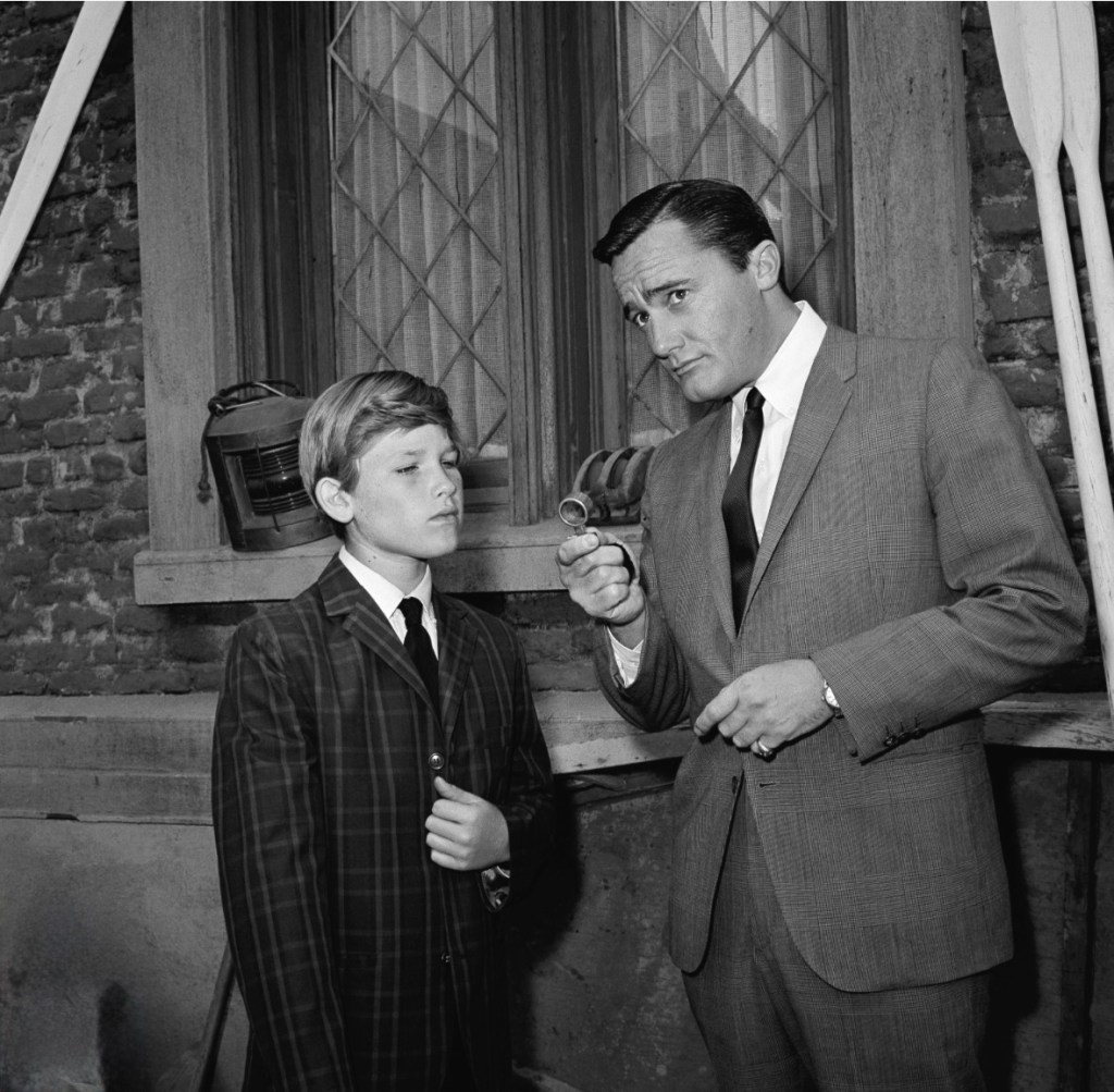 A serious-looking man in a suit is showing a small object to a young boy dressed in a plaid suit, both standing in front of a window with a brick wall behind them.