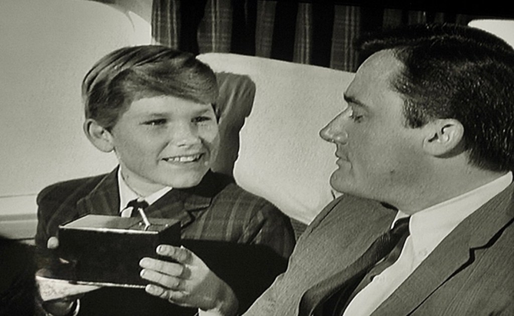 A young boy with short hair smiles while holding a small box, engaging with an adult man in a suit, seated in an airplane cabin.