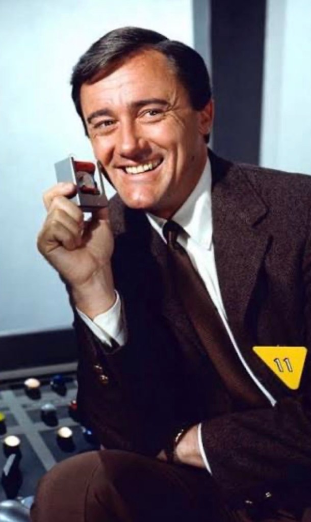 A smiling man in a brown suit holding a small device to his ear, sitting in front of control panel buttons.