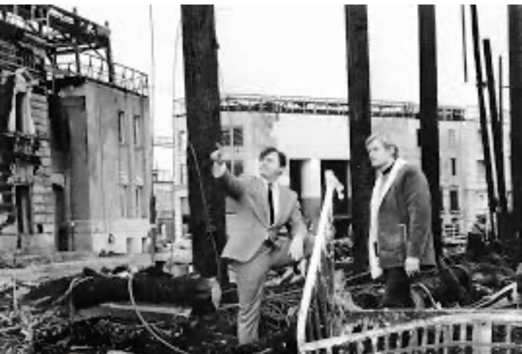 Two men examining an industrial site with visible damage and debris, under a cloudy sky.