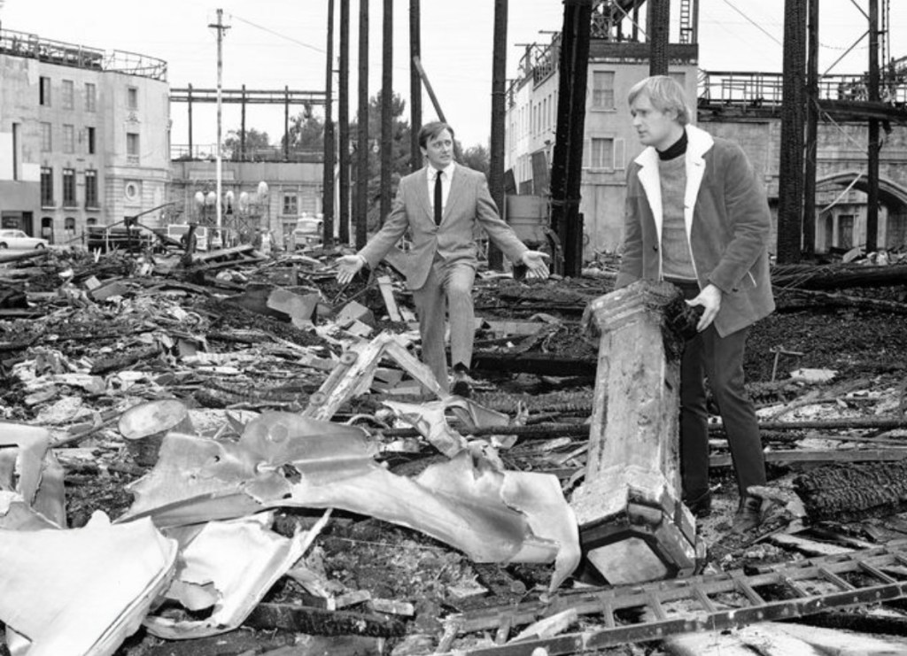 Two men standing amidst rubble and debris in a black and white scene, with damaged structures and burnt materials surrounding them.
