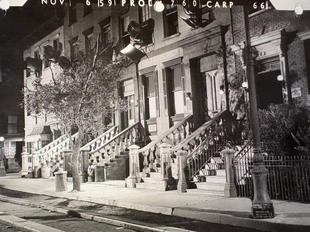 Black and white photograph of a city street featuring brownstone buildings with prominent staircases, street lamps, and a tree. The scene captures a nostalgic urban atmosphere.