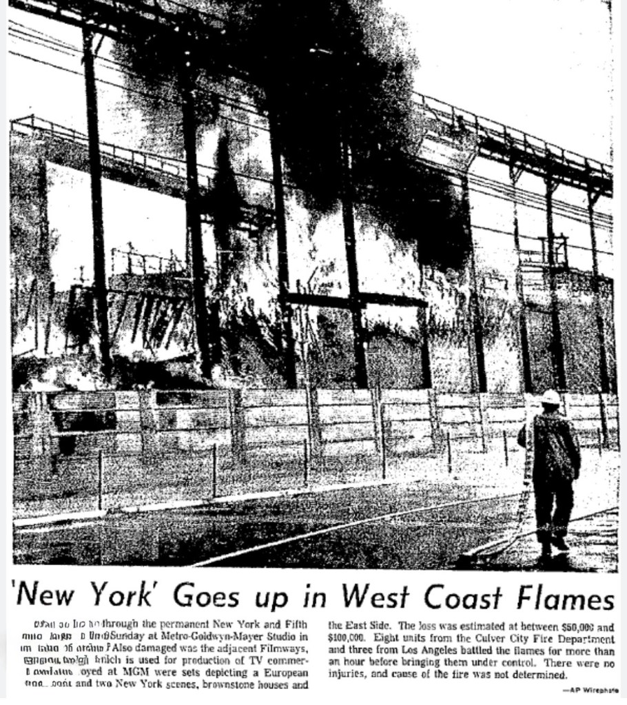 A black and white news image depicting a large fire engulfing a building structure with flames and smoke billowing, accompanied by firefighters working to control the blaze.